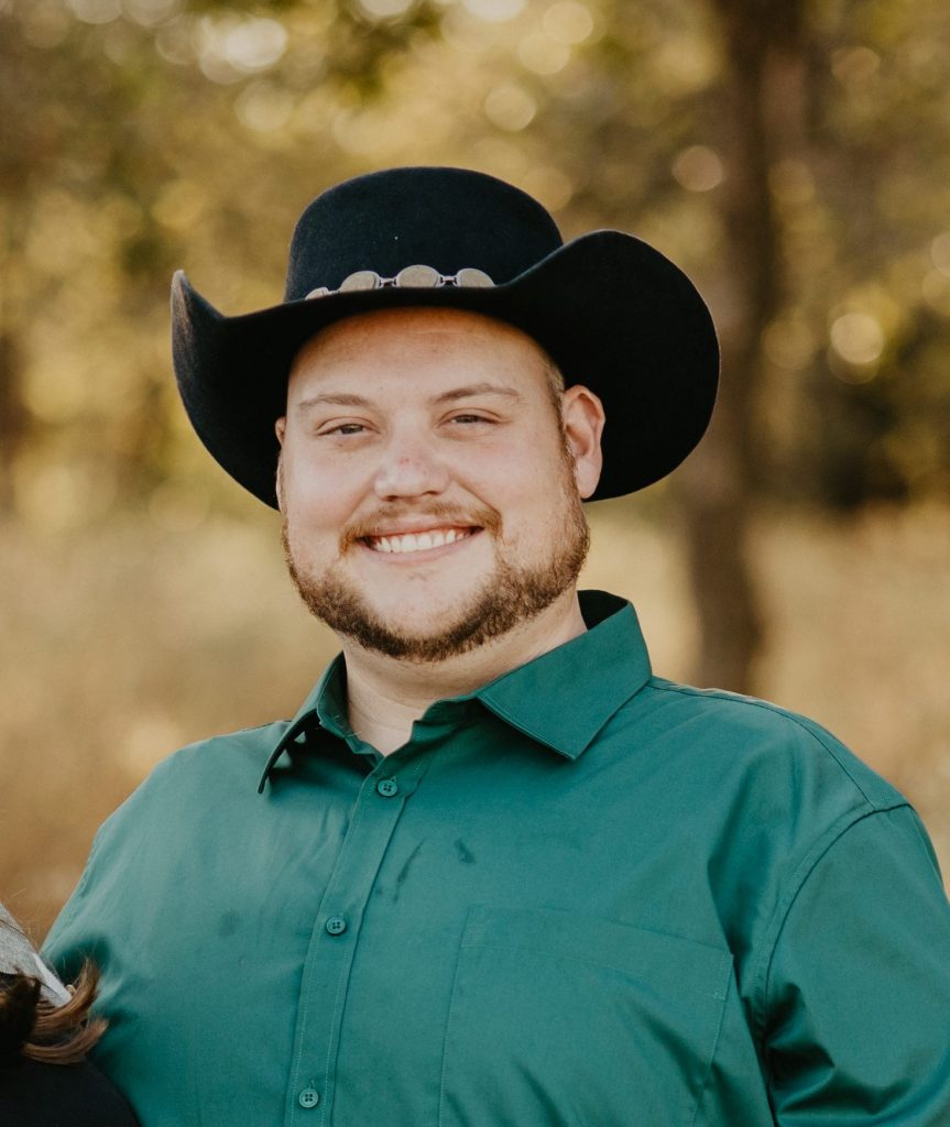 Dylan in a black cowboy hat and green button-up shirt with autumn trees in the background